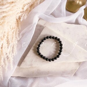 May include: A black beaded bracelet on a white marble surface. The bracelet is made of round, black beads.