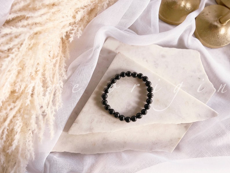 May include: A black beaded bracelet on a white marble surface. The bracelet is made of round, black beads.