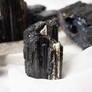 May include: Black tourmaline crystal with white streaks, a popular stone for grounding and protection. The crystal is sitting on a white fabric surface.