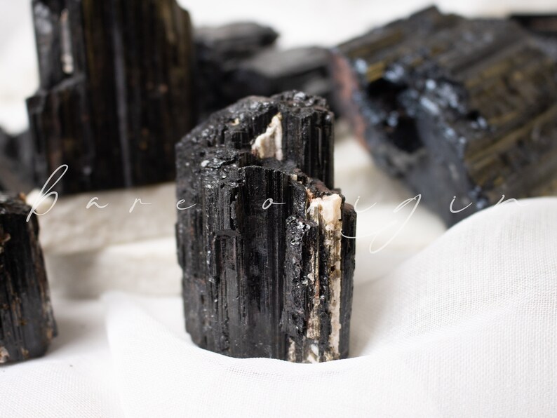May include: Black tourmaline crystal with white streaks, a popular stone for grounding and protection. The crystal is sitting on a white fabric surface.