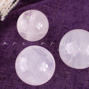 May include: Three clear quartz crystal spheres on a purple textured surface. The spheres have a translucent appearance with internal inclusions and subtle rainbow iridescence. The text "bare or virgin" is written in cursive across the image.