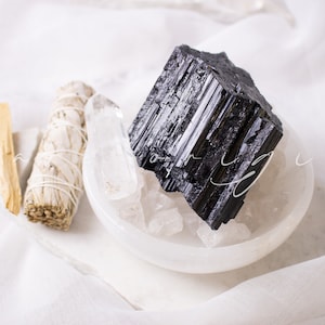 May include: A black tourmaline crystal in a white bowl with white sage and palo santo sticks. The crystals are on a white fabric background.