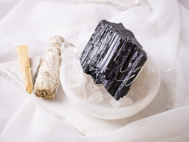 May include: A black tourmaline crystal in a white bowl with white sage and palo santo sticks. The crystals are on a white fabric background.