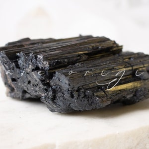 May include: A large, black, rough, and jagged piece of black tourmaline crystal. The crystal has a striated texture and a dark, almost metallic sheen.