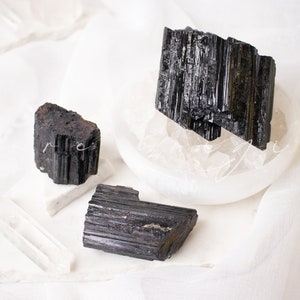 May include: Three black tourmaline crystals on a white surface. The crystals are raw and have a striated texture.