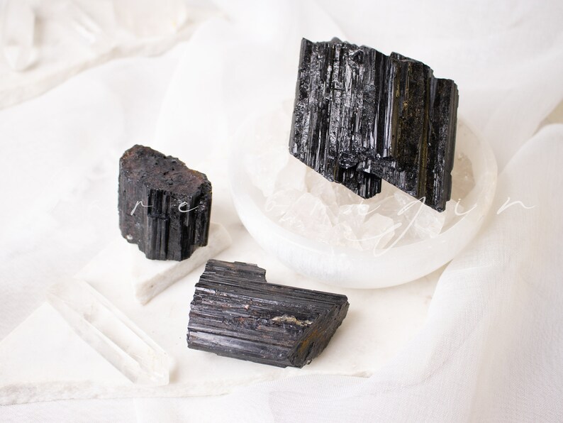 May include: Three black tourmaline crystals on a white surface. The crystals are raw and have a striated texture.