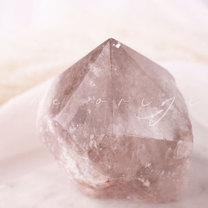 May include: A smoky quartz crystal point with a faceted top. The crystal is a translucent, light brown color and sits on a white surface. The word "bare origin" is written in cursive across the image.