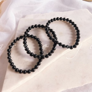 May include: Three black beaded bracelets stacked on a white surface. The bracelets are made of round, smooth beads.