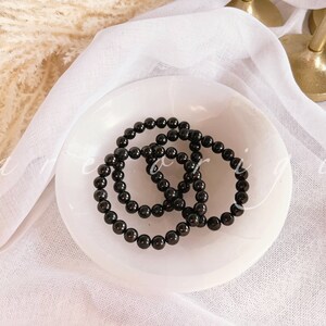 May include: Three black beaded bracelets stacked on a white ceramic dish. The bracelets are made of round, smooth beads.