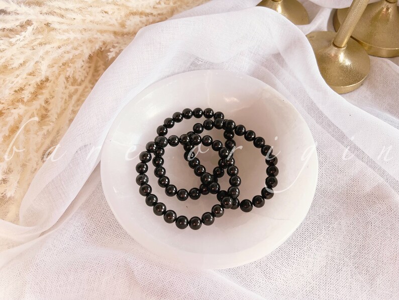 May include: Three black beaded bracelets stacked on a white ceramic dish. The bracelets are made of round, smooth beads.