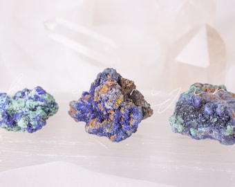 May include: Three rough, natural, blue and green azurite and malachite crystal specimens on a white selenite slab.
