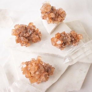 May include: Four clusters of brown selenite crystals on white marble surfaces. The crystals are arranged in a symmetrical pattern.