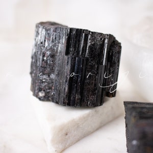 May include: A black tourmaline crystal with a rough, natural texture. The crystal is sitting on a white surface.