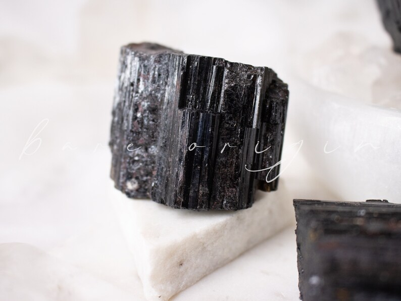 May include: A black tourmaline crystal with a rough, natural texture. The crystal is sitting on a white surface.