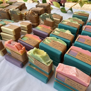 May include: A selection of handmade soaps in shades of green, pink, purple, and beige. Each bar is wrapped with a brown paper label. The soaps are arranged on a white surface, creating a visually appealing display of artisan products.