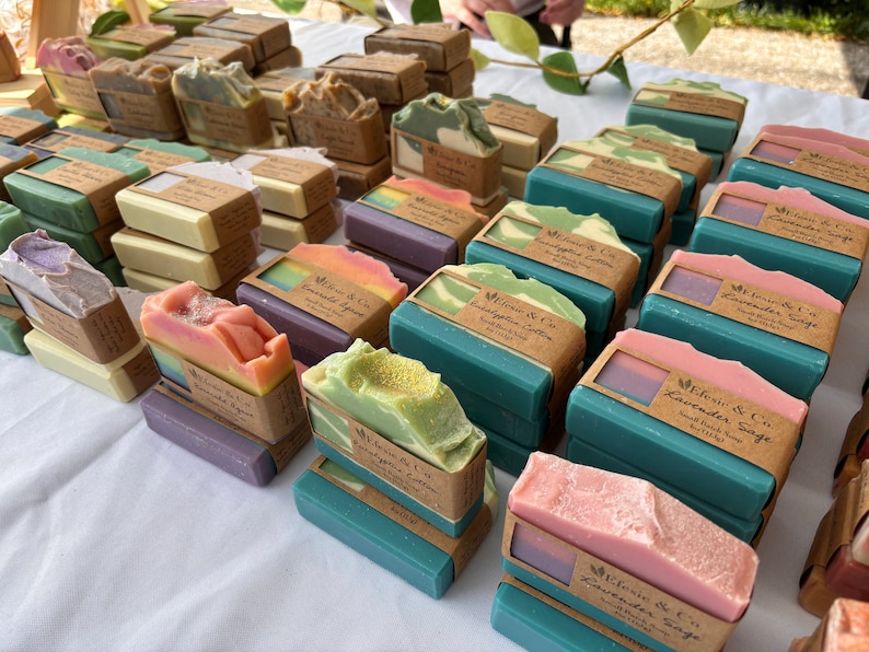 May include: A selection of handmade soaps in shades of green, pink, purple, and beige. Each bar is wrapped with a brown paper label. The soaps are arranged on a white surface, creating a visually appealing display of artisan products.