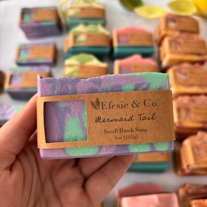May include: A close-up of a handmade soap bar with purple, green, and white swirls. The soap is labelled "Mermaid Tail" and is 113g. Other soap bars in various colours are in the background.