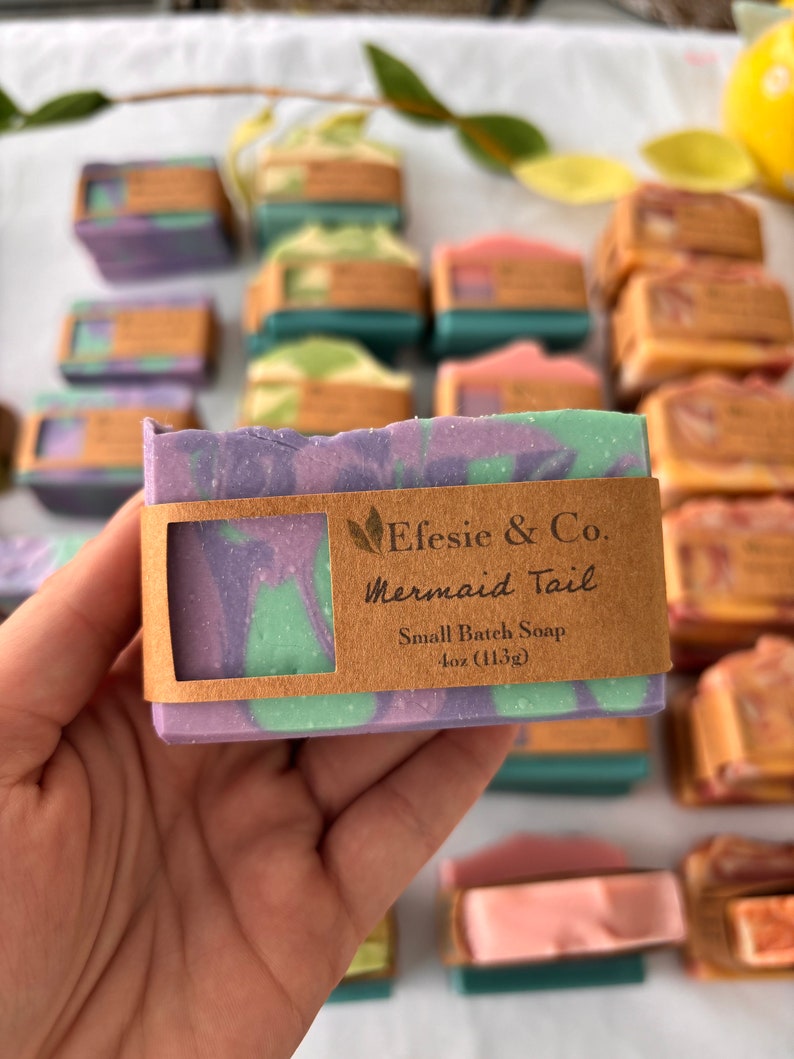 May include: A close-up of a handmade soap bar with purple, green, and white swirls. The soap is labelled "Mermaid Tail" and is 113g. Other soap bars in various colours are in the background.