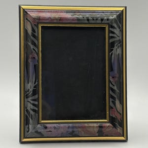 Puede incluir: Marco de fotos rectangular con interior negro. El marco tiene una base negra con un estampado floral en tonos morado, rosa y blanco. Un fino borde dorado bordea los bordes interior y exterior del marco.