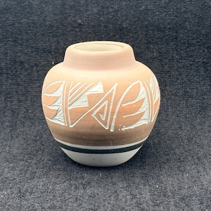 May include: A small, round, hand-painted pottery vase with a light peach top and a brown base. The vase features white and peach geometric designs. A black and white stripe encircles the base.