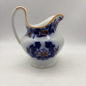 May include: White ceramic pitcher with a curved handle and spout. The pitcher is decorated with blue and orange floral patterns. The rim and handle are accented with orange trim. The pitcher has a bulbous body and a small foot.