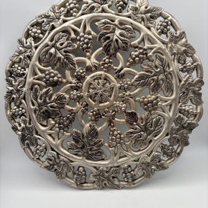 May include: Silver-colored, circular decorative plate with a grape and leaf design. The plate features a detailed, openwork pattern of grape clusters and leaves, creating a textured, elegant appearance. The plate is approximately 12 inches in diameter.