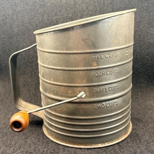 May include: Vintage metal flour sifter with a handle and wooden knob. The sifter has measurements for two, three, four, and five cups. The sifter is a dark, aged metal with visible wear and tear, suggesting it is an antique kitchen tool.