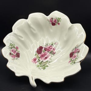 May include: A white, leaf-shaped ceramic serving bowl with a gold rim. The bowl is decorated with clusters of pink and burgundy roses and green leaves. The bowl has a decorative handle.