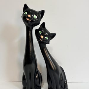 May include: Two black cat figurines with elongated necks and bodies. Each cat features bright green eyes, a small pink nose, and a subtle smile. The taller cat stands to the left, while the slightly shorter cat is positioned to the right.