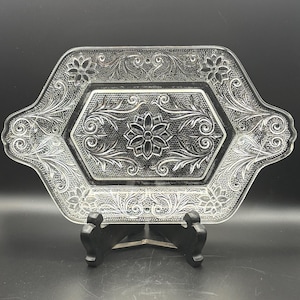 May include: Clear glass serving tray with an ornate floral and scroll design. The tray has a hexagonal shape with handles and is supported by a black stand. The intricate details of the embossed pattern are visible.