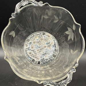 May include: Clear glass flower arranging bowl with etched leaf designs and two handles. The bowl has a round center with multiple holes for arranging flowers. The rim has a scalloped edge.