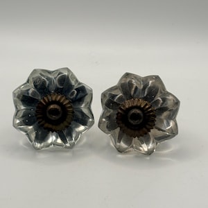 May include: Two clear glass flower-shaped cabinet knobs with a bronze-colored center. The knobs have a faceted design, catching the light. These decorative knobs are suitable for furniture restoration or adding a vintage touch to cabinets and drawers.