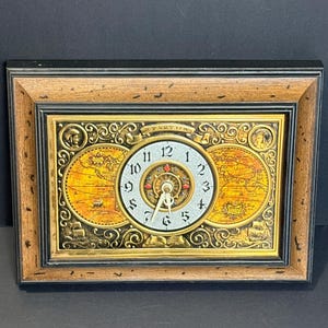 May include: A rectangular clock with a wooden frame. The clock face is surrounded by a gold-colored design with a world map on each side. The clock face has black numbers and hands.