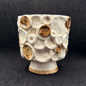 May include: Cream-colored ceramic planter with a textured, bubble-like design and a pedestal base. The raised circles are accented with brown, creating a contrast against the off-white background. The planter has a wide opening and is suitable for small plants.