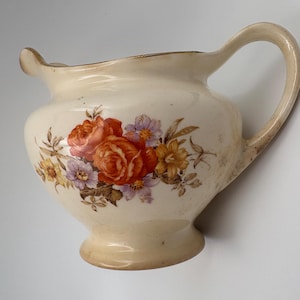 May include: Cream-coloured ceramic creamer with a floral design featuring orange roses, purple and yellow flowers. The creamer has a curved handle and a spout, with a gold trim around the rim. The base is a light beige colour.
