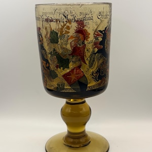 May include: A vintage amber-colored glass goblet with a detailed painted scene of figures and text. The goblet has a round base, a bulbous stem, and a wide rim. The painted scene features various characters in vibrant colors.