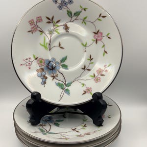 May include: A set of white porcelain saucers with a silver rim and floral design. The floral pattern includes pink, blue, and green flowers and leaves. The saucers are stacked on a black stand.
