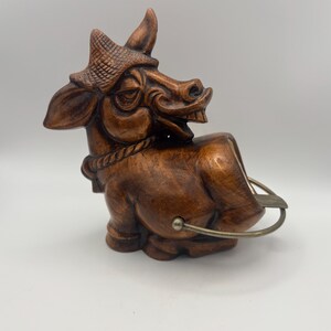 May include: A brown, carved wooden figurine of a bull wearing a hat and rope around its neck. The bull is in a seated position with a metal ashtray attached to its side. The figurine has a detailed, expressive face and is likely vintage.