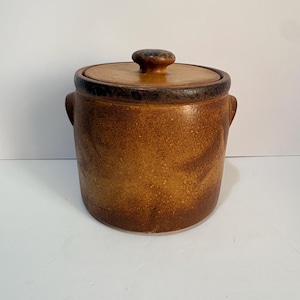 May include: A cylindrical stoneware crock with a speckled brown glaze and a matching lid. The container features two small side handles and a knob on the center of the lid. The finish transitions from a lighter golden brown to a darker brown rim.