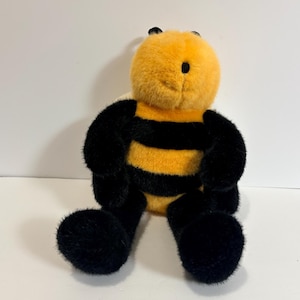 May include: A plush bee toy with a yellow head and body, and black stripes. The bee has black antennae, a black eye, and black legs. The toy is sitting on a white surface.