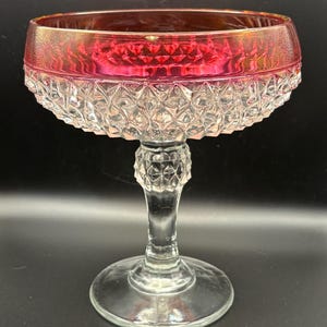 May include: A clear glass champagne coupe with a ruby red rim. The bowl has a diamond pattern, and the stem and base are also clear glass. The coupe is designed for serving champagne or other sparkling wines.