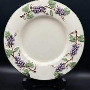 Puede incluir: Plato de cerámica blanquecino con borde verde y diseño de racimos de uvas. El plato presenta uvas moradas en relieve, hojas verdes y vides marrones. El plato se exhibe sobre un soporte negro.