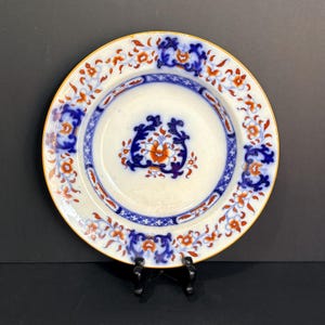 May include: A white ceramic plate with a detailed floral design in orange and blue. The plate has a gold rim and a central floral motif. The design includes a blue border with an intricate pattern. The plate is displayed on a black stand.