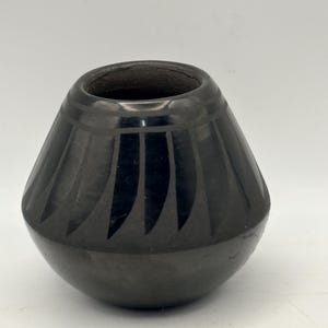 May include: A black ceramic pot with a rounded body and a wide opening. The exterior features a polished finish with a matte black band of vertical, petal-like shapes encircling the upper portion, separated by a thin horizontal line.