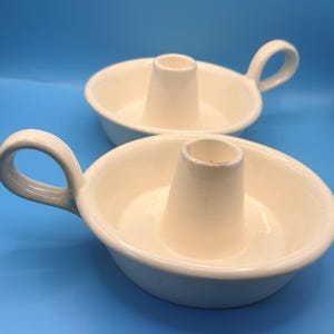 Puede incluir: Dos candeleros de cerámica blanquecina con asas. Cada uno tiene una base circular, un cono central para la vela y un asa curvada. El fondo es azul liso.