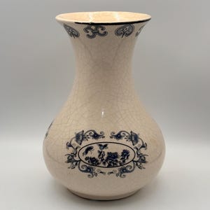 Puede incluir: Jarrón de cerámica color crema con acabado craquelado y diseños florales azules. El jarrón tiene un cuerpo bulboso y un cuello acampanado, con un diseño ovalado floral azul en la parte delantera. El borde está adornado con un patrón azul.