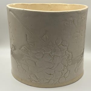 Puede incluir: Un recipiente cilíndrico de cerámica blanquecina con patrones florales y de hojas en relieve. El recipiente tiene una abertura ligeramente ovalada y una base clara. El diseño está en relieve, creando una superficie texturizada. La estética general es rústica y vintage.