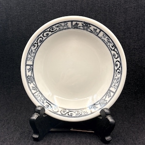 May include: A white ceramic bowl with a decorative blue and black border. The bowl is displayed on a black stand against a dark background. The border features a repeating pattern of stylized floral and scroll designs.