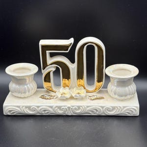 Pode incluir: Centro de mesa de cerâmica para uma celebração de 50 anos. O número "50" é dourado e branco, ladeado por dois pequenos castiçais. A base é decorada com detalhes florais.