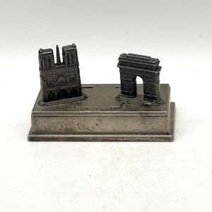 Könnte beinhalten: Eine silberfarbene Metall-Souvenirfigur mit Miniaturnachbildungen der Kathedrale Notre-Dame und des Arc de Triomphe auf einem rechteckigen Sockel. Das Stück hat eine verwitterte, dunkelgraue Oberfläche mit detaillierten architektonischen Strukturen.